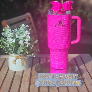May include: Pink stainless steel tumbler with a pink handle and a pink bow on top. The tumbler is covered in rhinestones and has the word "Stanley" printed on it. The tumbler is sitting on a wooden table with a sticker that says "Hustle Monkey Custom Creations".