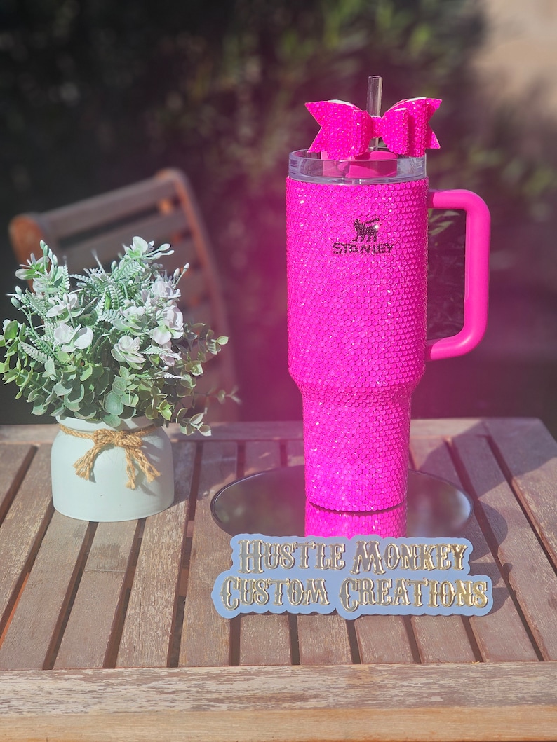 May include: Pink stainless steel tumbler with a pink handle and a pink bow on top. The tumbler is covered in rhinestones and has the word "Stanley" printed on it. The tumbler is sitting on a wooden table with a sticker that says "Hustle Monkey Custom Creations".