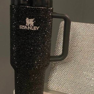 Puede incluir: Taza de viaje Stanley negra con un dise&ntilde;o de pedrer&iacute;a negra y un lazo negro en la tapa. La taza tiene un asa negra y la palabra "STANLEY" est&aacute; escrita en la parte delantera. Tres bol&iacute;grafos de pedrer&iacute;a negra est&aacute;n sobre una superficie blanca.