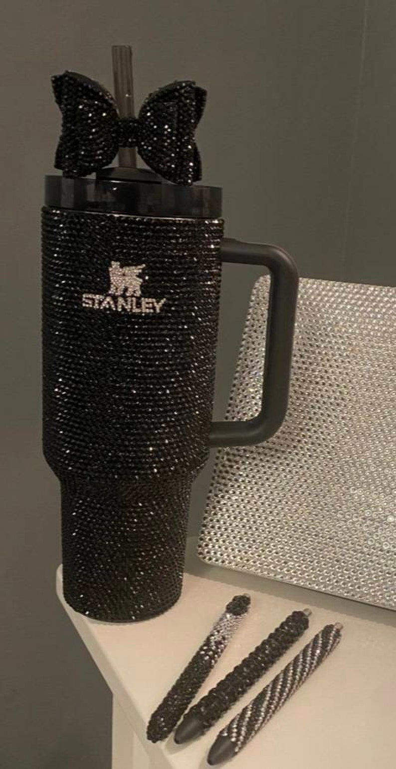 Puede incluir: Taza de viaje Stanley negra con un dise&ntilde;o de pedrer&iacute;a negra y un lazo negro en la tapa. La taza tiene un asa negra y la palabra "STANLEY" est&aacute; escrita en la parte delantera. Tres bol&iacute;grafos de pedrer&iacute;a negra est&aacute;n sobre una superficie blanca.
