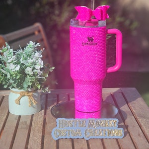 May include: A pink Stanley tumbler with a pink handle and a pink bow on top. The tumbler is covered in rhinestones and has the Stanley logo on the side. The tumbler is sitting on a wooden table with a sign that says "Hustle Monkey Custom Creations".
