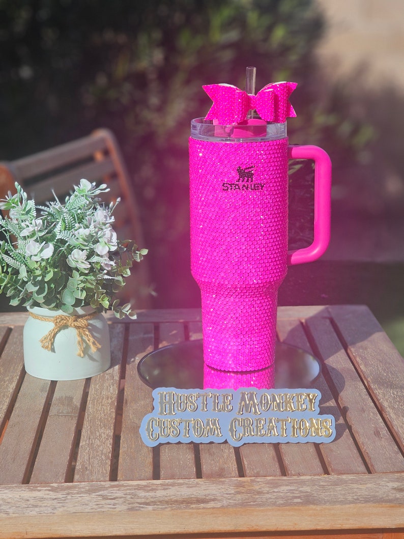 May include: A pink Stanley tumbler with a pink handle and a pink bow on top. The tumbler is covered in rhinestones and has the Stanley logo on the side. The tumbler is sitting on a wooden table with a sign that says "Hustle Monkey Custom Creations".