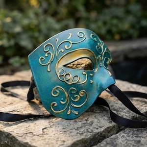 May include: A teal masquerade mask featuring raised gold filigree scrollwork patterns across the surface. The mask has a single eye opening and includes long black satin ribbons attached to the sides for securing the item to the face.