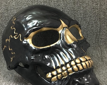 May include: A black plastic skull mask with gold accents. The mask has a detailed skull design with gold teeth and eye sockets.