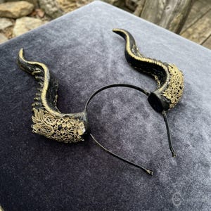 May include: A thin black metal headband featuring two curved, textured black horns. Each horn is accented with intricate gold-colored lace detailing applied to the base and along the outer ridges, creating a decorative headpiece accessory.