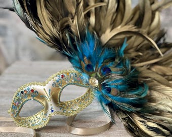 May include: A gold masquerade mask with blue and gold feathers. The mask has a delicate design with rhinestones and glitter.