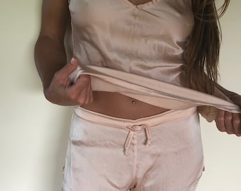 May include: A pale pink satin pajama set consisting of a sleeveless camisole top with a V-neckline and matching shorts featuring an elasticized waistband with a decorative drawstring tie. The fabric has a smooth, lustrous finish throughout.