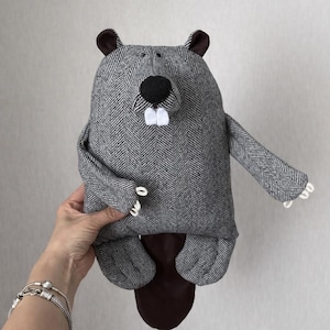 May include: A gray and white stuffed beaver toy with a herringbone pattern. The beaver has a black nose, brown ears, and two white teeth. The beaver's tail is brown. The toy is being held by a person.