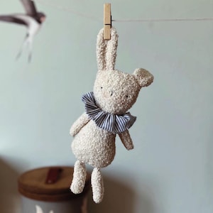 May include: A beige bunny plush toy with a blue and white striped collar is suspended from a clothesline by a wooden clothespin. The bunny has long ears and small arms and legs. A gray and brown storage container with white bird silhouettes is in the background.