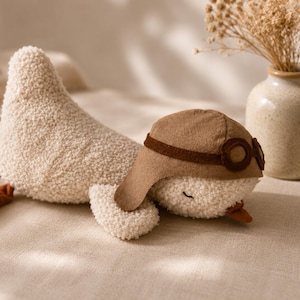 Puede incluir: Un peluche de ganso color crema con un cuerpo de tela texturizada tipo sherpa. El ganso lleva un gorro de aviador de fieltro marrón con gafas incorporadas, tiene patas de tela color óxido y un pequeño pico naranja, en posición de reposo.