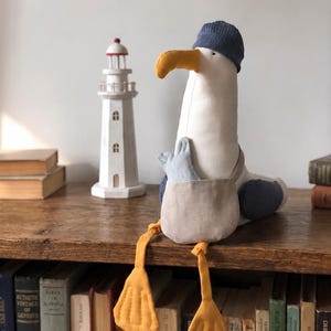 May include: A plush seagull toy with a blue knit cap, yellow beak and feet, and a tan pouch. The seagull is sitting on a wooden shelf next to a white lighthouse model and a stack of books. The seagull's feet are approximately 4 inches long.
