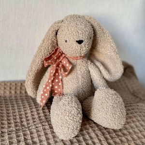 Könnte beinhalten: Ein Plüschkaninchen mit langen Schlappohren und einer hellbraunen, strukturierten Außenseite. Das Stofftier hat eine rosa Schleife mit Punkten um den Hals und sitzt auf einer strukturierten Decke.