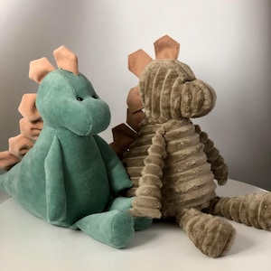 Może przedstawiać: Dwie pluszowe zabawki dinozaurów. Jedna jest miękko zielona z kolcami w kolorze brzoskwiniowym, druga ma brązową fakturę z kolcami w kolorze brzoskwiniowym. Oba siedzą na białej powierzchni.