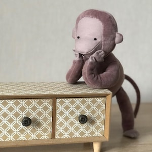 Puede incluir: Un peluche de mono de color malva descansa sobre una pequeña cómoda de madera. La cómoda tiene tres cajones con un patrón geométrico en blanco y beige, y pomos negros. El mono está apoyado en la cómoda con las manos en la cara.