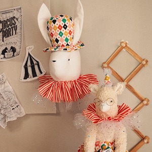 Puede incluir: Dos figuras de peluche con temática de circo, un conejo y una llama, se exhiben sobre un fondo neutro. El conejo lleva un sombrero estampado y un cuello a rayas rojas y blancas. La llama lleva gafas y sostiene una entrada de circo.