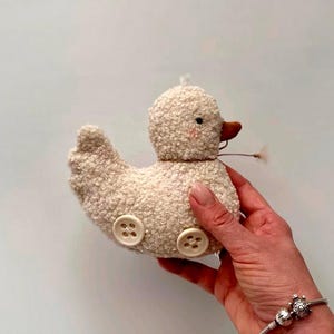 Pode incluir: Um brinquedo de pato de pelúcia em tecido creme com rodas de botão. O pato tem um bico marrom, bochechas rosadas e uma cabeça pequena e fofa. Uma pequena flor seca está presa ao bico. O brinquedo foi projetado para se parecer com um carro vintage.