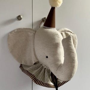 Könnte beinhalten: Wanddekoration in Form eines Elefantenkopfes aus beigem Stoff, mit braunem Partyhut und weißem Bommel. Der Elefant hat große Ohren und einen Rüschenkragen mit braunen Streifen.