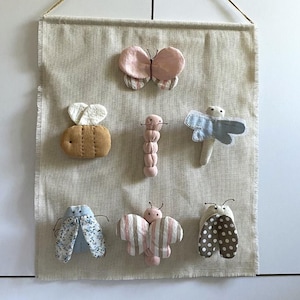 以下が含まれることがあります： ベージュ色の布製の壁掛けで、さまざまな布製の昆虫の装飾が施されています。昆虫には、ミツバチ、蝶、イモムシ、トンボ、蛾が含まれます。昆虫はピンク、青、茶色、白の色合いです。壁掛けは天然のロープで吊り下げられています。