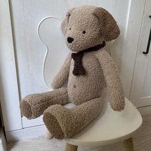 May include: A soft toy dog in shades of brown, with darker brown ears and paws, and a brown scarf. The dog is seated on a white chair with wooden legs. The toy has a black nose and button eyes.