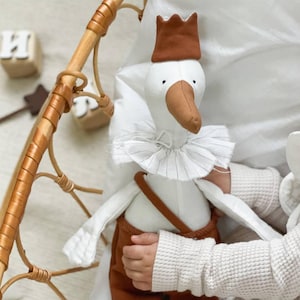 May include: A plush goose doll wearing a brown crown, pants, and suspenders, with a white ruffled collar. The doll has a brown beak and feet. The doll is being held by a child in a light-colored sweater.