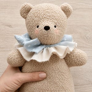 May include: A beige crocheted teddy bear with a textured surface, featuring small black eyes, a black nose, and soft pink cheeks. The bear wears a layered ruffled collar made of light blue fabric with white polka dots and a plain cream-colored fabric.