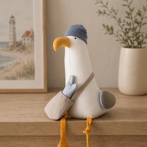 May include: A plush seagull toy with a white body, a yellow beak, and long yellow legs. The seagull wears a blue ribbed knit beanie and carries a light beige crossbody bag containing a small blue mitten. A grey wing is visible on its side.