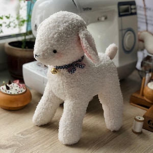 Può includere: Un giocattolo fatto a mano a forma di agnello di peluche bianco con una consistenza riccia. L'agnello ha una piccola campana su un collare a fantasia blu e bianco. L'agnello ha accenti rosa sulle orecchie e sulle guance. Sullo sfondo c'è una macchina da cucire.