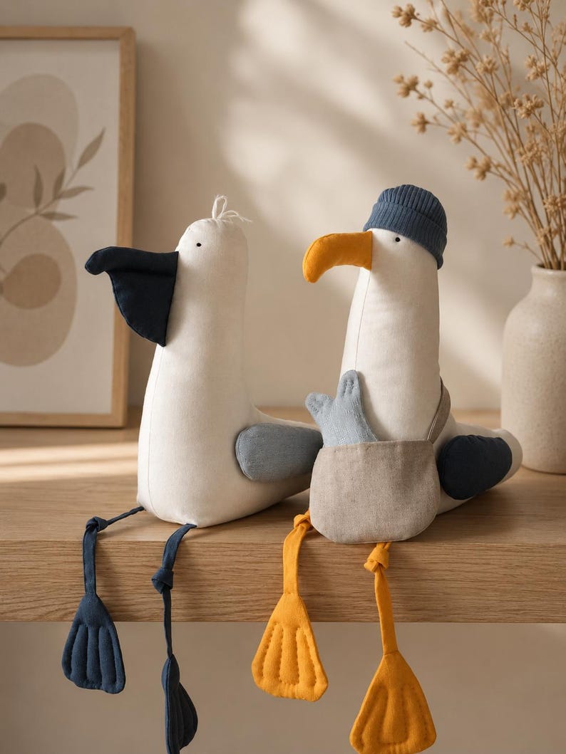 May include: Two plush seagull toys with off-white bodies. The left seagull has a dark navy beak, wings, and feet. The right seagull wears a blue ribbed beanie, has a yellow beak, yellow feet, and carries a small beige satchel with a light blue fish.