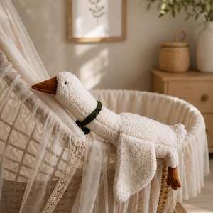 Puede incluir: Un juguete de peluche con forma de ganso hecho de tela blanca texturizada con pico y patas marrones. El ganso lleva un cuello tejido de color verde oscuro alrededor del cuello y está colocado dentro de un moisés tejido cubierto con tela blanca.