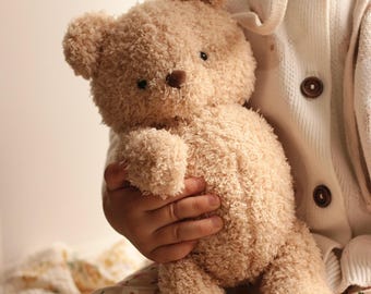 Puede incluir: Un oso de peluche marrón claro con un exterior texturizado y difuso. El oso tiene una pequeña nariz marrón y ojos negros. El oso está siendo sostenido por una persona que lleva una rebeca blanca con botones marrones. El fondo es suave y borroso.