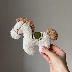 Può includere: Un giocattolo a forma di cavallo bianco in peluche, con criniera e coda marroni e una sella verde. Il giocattolo è tenuto in mano su uno sfondo grigio. Il cavallo ha un design semplice, con un viso cucito e una piccola briglia.