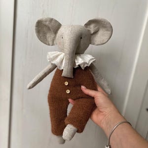 Puede incluir: Una muñeca de elefante de peluche hecha a mano con orejas grandes, una trompa larga y un cuello con volantes. El elefante lleva pantalones marrones con botones de madera. La muñeca está hecha de tela suave de tonos neutros.