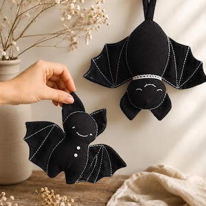 Può includere: Due ornamenti a forma di pipistrello in tessuto nero con dettagli delle ali cuciti in bianco. Un pipistrello è appeso a un piolo di legno e presenta un colletto in pizzo bianco. L'altro è tenuto in mano, mostrando due bottoncini bianchi.