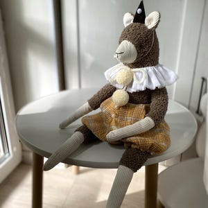 May include: A brown plush animal doll with a party hat, white ruffled collar, and yellow plaid dress. The doll has long, striped arms and legs, and is sitting on a small, round, white table. The doll's ears are white.