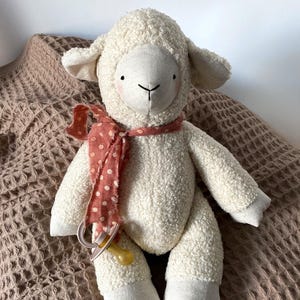 Puede incluir: Una muñeca de peluche de cordero blanco con cara, orejas y pies color crema. El cordero tiene una bufanda roja con lunares y un chupete. La muñeca está sobre una manta marrón texturizada.