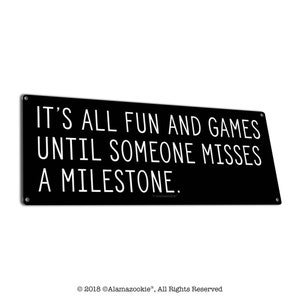 May include: Black and white sign with the text "IT'S ALL FUN AND GAMES UNTIL SOMEONE MISSES A MILESTONE." The sign has a white border and a small copyright symbol with the text "Alamazookie" in the bottom right corner.