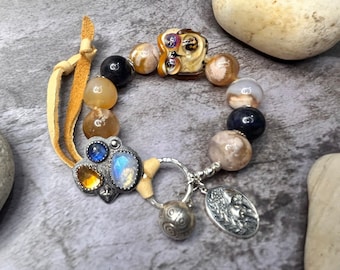 May include: A brown leather bracelet with agate, blue, and glass beads. The bracelet has a silver charm with a moonstone, sapphire, and citrine stones. The charm is attached to a silver ring with a silver charm in the shape of a ladybug.