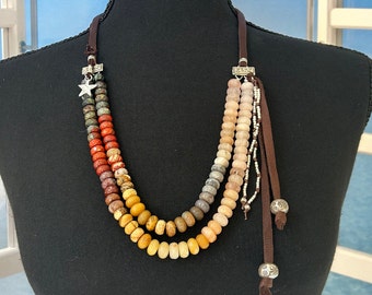 Puede incluir: Un collar de múltiples hilos con cordones de cuero marrón y detalles plateados. El collar presenta una variedad de cuentas redondas de piedras preciosas de colores en tonos rojos, naranjas, amarillos y blancos.