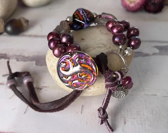 May include: A multi-strand bracelet featuring deep purple pearls, a large circular button with a metallic silver lizard motif over an iridescent orange and purple background, and brown leather cords with small beads and a silver-toned charm.