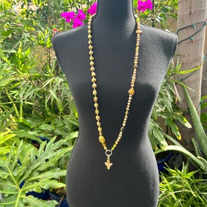 May include: A long gold and brown beaded necklace with a gold pendant. The necklace is displayed on a black mannequin.
