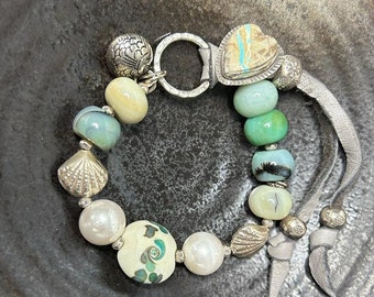 May include: A silver and turquoise beaded bracelet with a heart charm, shell charms, and a silver toggle clasp. The bracelet is made with a variety of beads, including turquoise, white, and green.