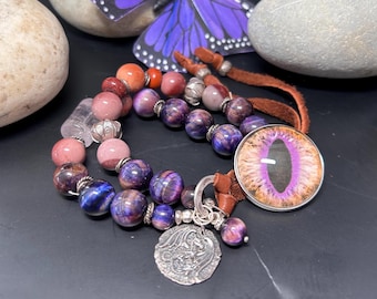 May include: A multi-strand bracelet with purple, pink, and red gemstone beads, a silver charm with a dragon design, and a purple eye charm.