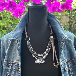 Può includere: Una collana multifilo con perline grigie e bianche, un anello d'argento e un ciondolo d'argento con un motivo floreale. La collana è su un manichino che indossa una giacca di jeans.