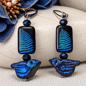 Puede incluir: Un par de pendientes colgantes con una cuenta rectangular negra con estampado de hoja de helecho azul, una cuenta pequeña facetada oscura y una cuenta azul texturizada con forma de pájaro. Enganchados a cierres de metal oscuro.