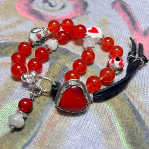 Könnte beinhalten: Ein rotes und weißes Perlenarmband mit einem Herz-Anhänger und einer schwarzen Lederschnur. Das Armband besteht aus roten Perlen, weißen Perlen und einem herzförmigen Anhänger. Der Anhänger ist aus Metall und hat einen roten Stein in der Mitte. Das Armband ist mit einer schwarzen Lederschnur gebunden.