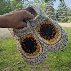 May include: A pair of handmade crochet slippers featuring a sunflower motif on the top. The slippers are constructed with a thick, cream-colored yarn base, while the sunflower design incorporates dark brown centers surrounded by golden yellow petals.