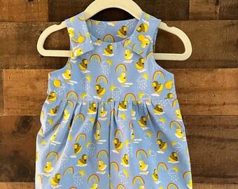 May include: A light blue baby romper with a yellow duck and rainbow print. The romper has a Peter Pan collar and buttons on the shoulders.