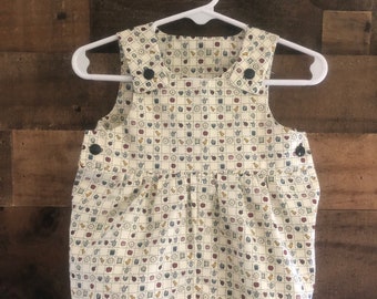 May include: A white baby onesie with a pattern of small, colorful illustrations of apples, birds, and other objects. The onesie has a bib-style top with straps and buttons.