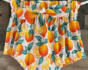 May include: Orange print baby bloomers with a white background and a green leaf pattern. The bloomers have a bow at the waist.