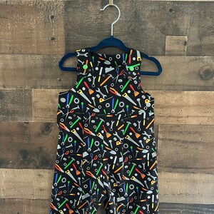May include: Black overalls with a colorful tool pattern. The overalls have adjustable straps and a button closure.